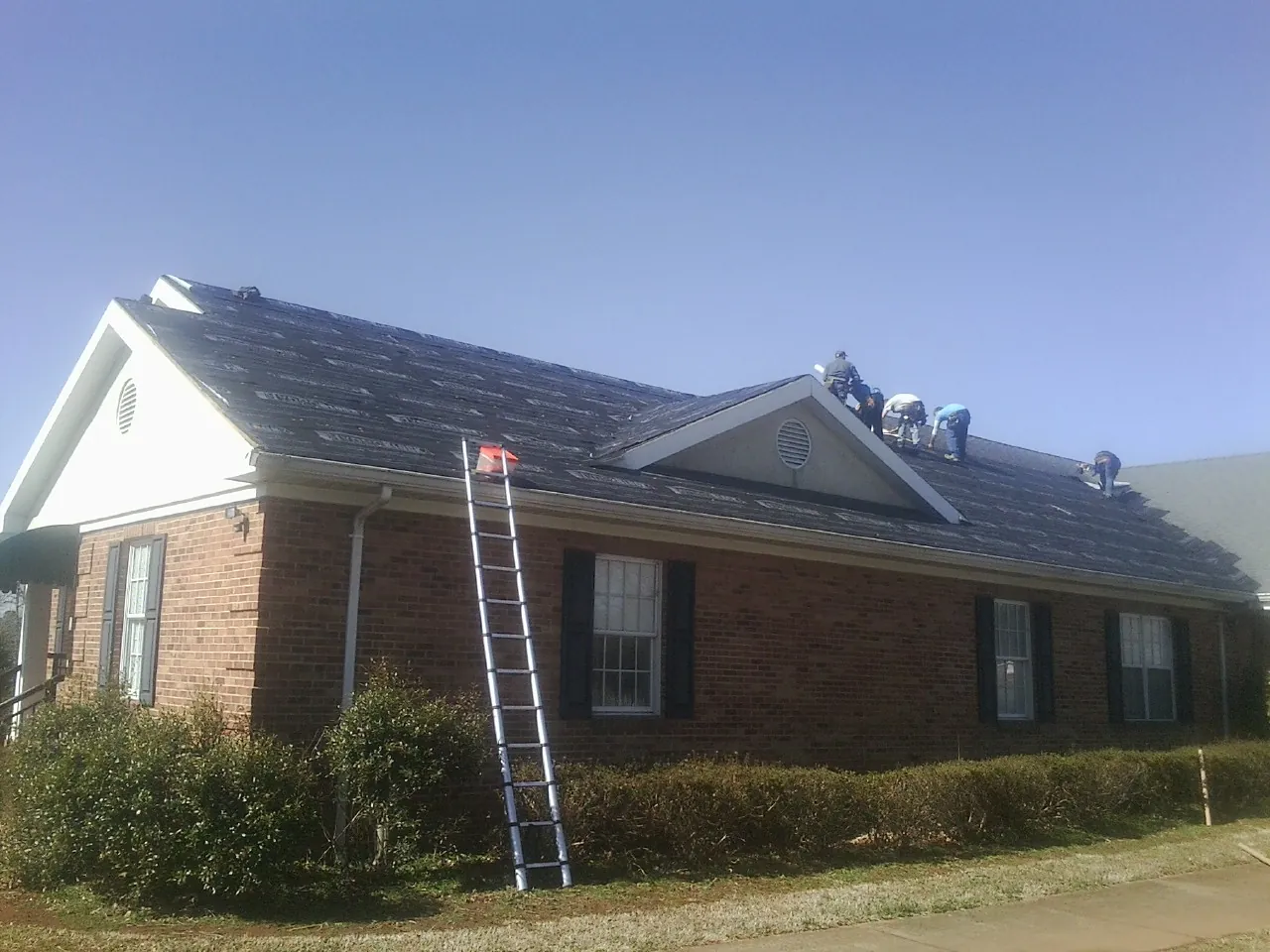 Crew tearing shingles off a brick ranch roof with a ladder set — Metal Roof Repair work by our crew in Byron