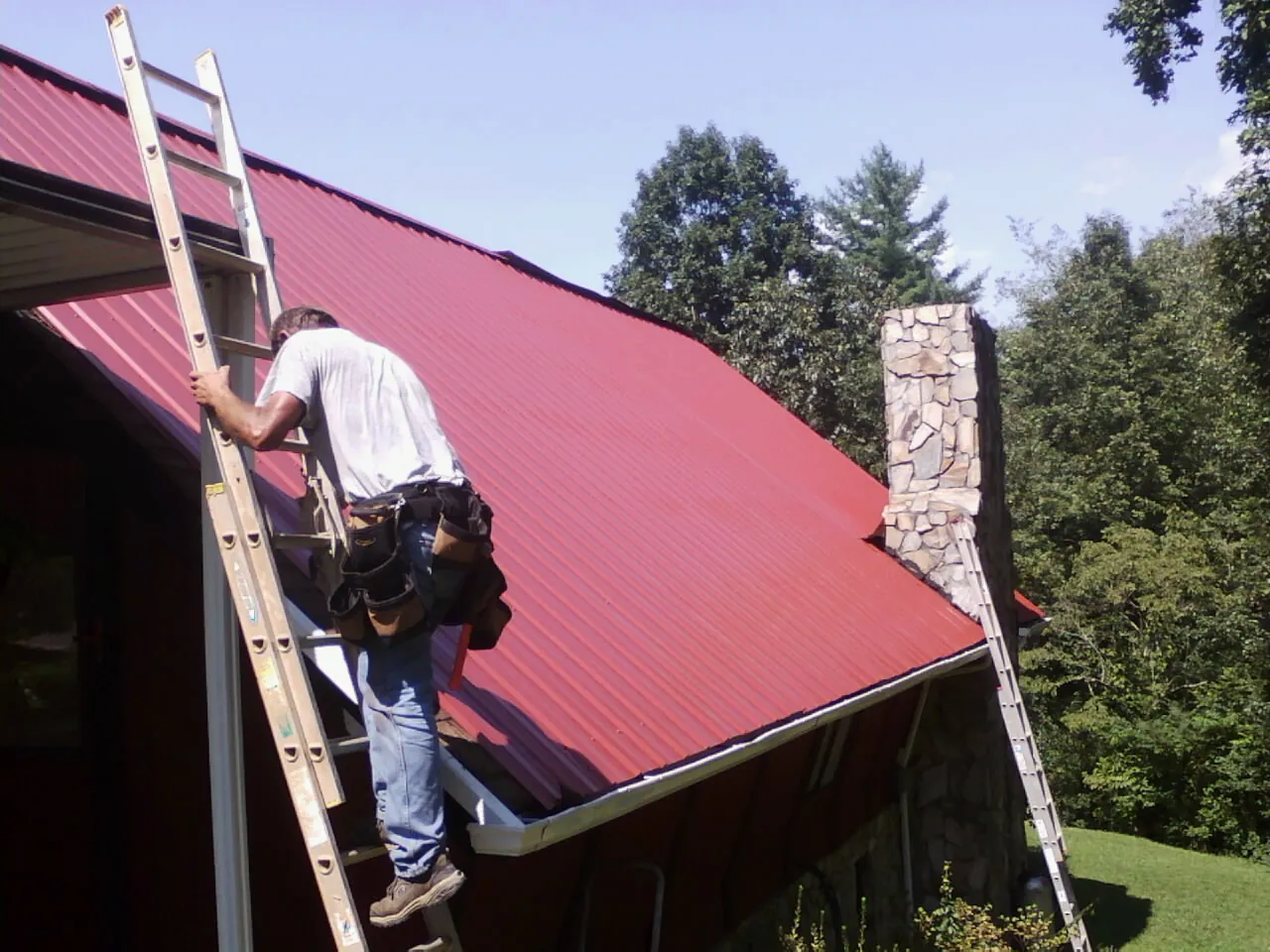 Roofer on a ladder beside a red metal roof and stone chimney — Soffit & Fascia Repair by Roofing Brothers in Byron